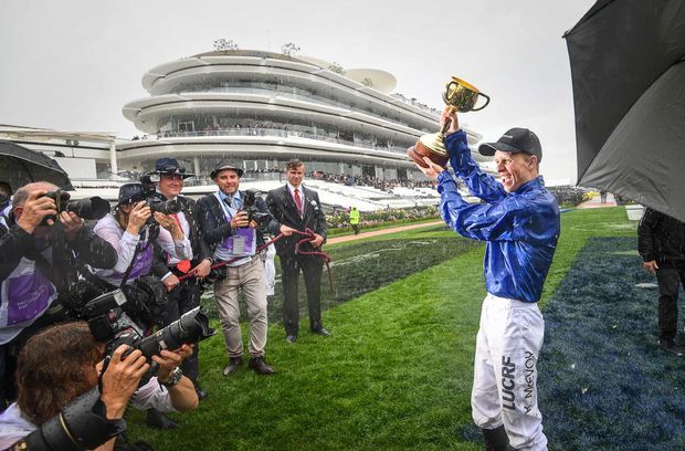 A press photograph on a rain-soaked horse-racing track, showing a jockey in blue racing silks holding a trophy while surrounded by photographers and media. A large multi-level grandstand is visible in the background.