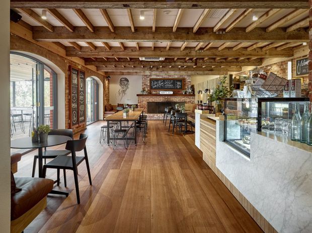 A warm, rustic café interior with exposed timber beams and rafters, wide timber flooring, brick walls, arched glazed doors, and industrial-style black dining furniture. A marble-faced service counter with glass display cases occupies the foreground, while a fireplace, chalkboard, and café seating are visible toward the rear. The space is bright and inviting, combining heritage character with contemporary hospitality fit-out.
