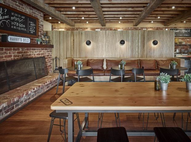 Rustic-industrial café or hospitality interior featuring a large communal timber table, leather banquette seating, black metal dining chairs and stools, exposed timber ceiling beams, vertical timber wall panelling, and a brick fireplace with chalkboard signage. The warm, earthy palette and layered timber and brick finishes create an inviting, casual atmosphere.