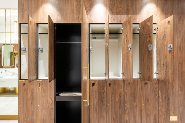 A wall of tall built-in timber wardrobes with multiple open doors, hanging rails, shelves, and lower storage compartments. The cabinetry is finished in rich mid-to-dark wood veneer with integrated lighting, and a bright bathroom or dressing area is partially visible through the opening at left.