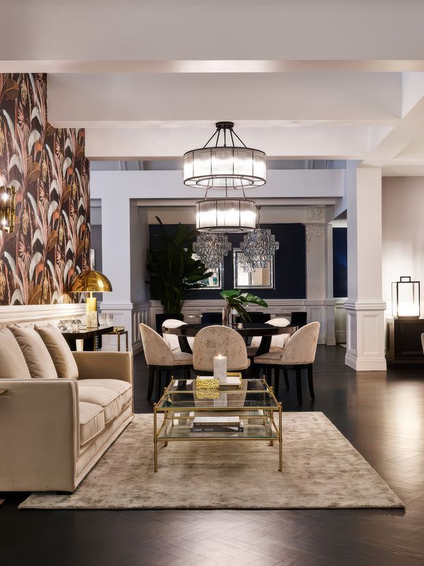 A refined open-plan living and dining area with a beige upholstered sofa, glass-and-brass coffee table, patterned feature wallpaper, large decorative pendant lights, and a dark blue dining-room feature wall. White architectural mouldings, columns, wainscoting, dark timber flooring, and elegant upholstered dining chairs create a luxurious, classic interior.