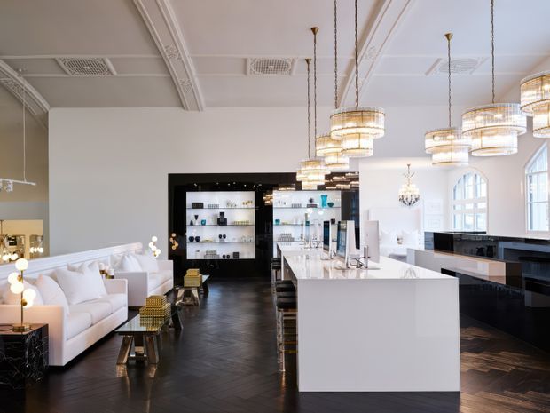 Luxurious open-plan kitchen and living area with a large white waterfall island, glossy black cabinetry, dark herringbone timber flooring, white upholstered seating, mirrored shelving, and multiple tiered crystal pendant lights. The room features high decorative ceilings, ornate mouldings, large arched windows, and a strong black-and-white palette with metallic accents.