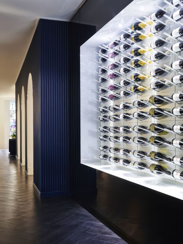 A dramatic, contemporary wine display wall occupies the right side of a narrow interior passage. Dozens of bottles are horizontally mounted in a bright, backlit white shelving system, contrasting with a dark navy-black fluted wall and dark herringbone timber flooring. A softly lit adjoining room is visible beyond the passage, with decorative arched openings along the left wall.