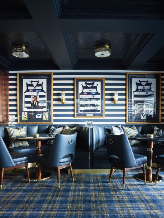 A richly detailed club-style lounge or sports-themed bar interior with navy and white striped walls, framed football memorabilia, deep blue leather banquette seating, upholstered dining chairs, small round tables, brass accents, and a dark coffered ceiling. A blue plaid carpet reinforces the refined collegiate and heritage character.