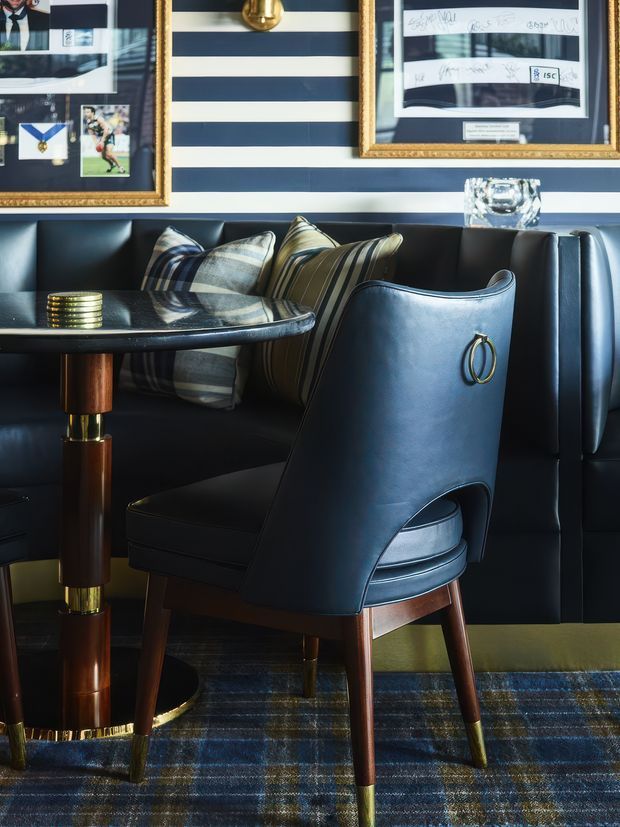 A richly styled dining nook or lounge corner with a dark navy upholstered banquette, matching leather dining chair, and a round black tabletop with polished wood-and-brass pedestal legs. The wall features bold horizontal navy-and-white stripes, framed artwork and photography in ornate gold frames, and a small brass wall light. Patterned cushions and a blue-toned plaid rug add texture, while metallic accents and dark timber create a sophisticated, tailored atmosphere.