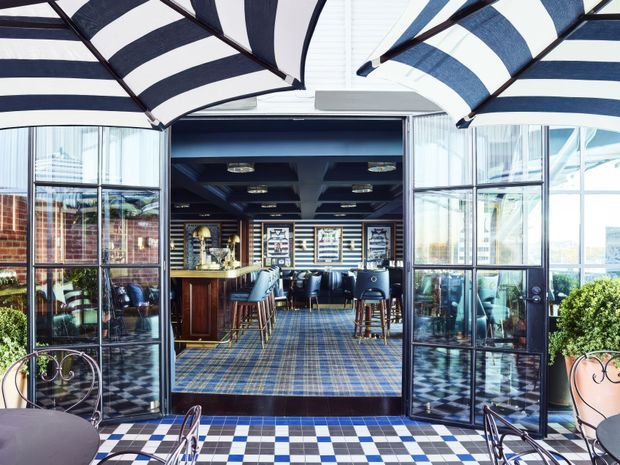 View from an outdoor terrace into a stylish commercial bar or restaurant through open black-framed glazed doors. The space features a striking blue, white and grey checkerboard terrace floor, blue patterned carpet, dark timber bar cabinetry, booth and bar seating, striped wall detailing, extensive glazing, and large navy-and-white striped awnings. Outdoor café tables and chairs, shrubs, and decorative ironwork frame the entrance.