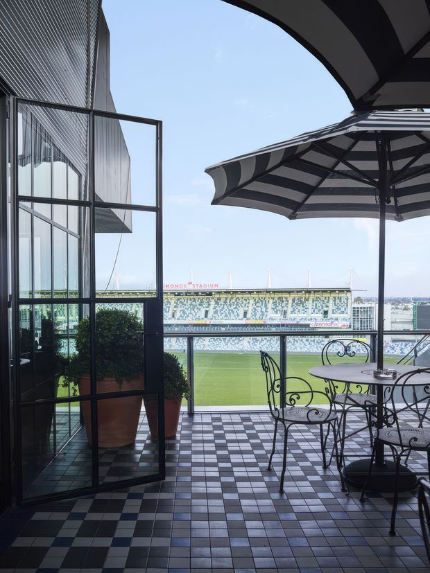 A covered outdoor terrace or balcony overlooking a sports stadium. The space has a black-and-white checkerboard tiled floor, black metal-framed glazing and doors, a glass balustrade, wrought-iron café table and chairs, large striped umbrellas, and potted plants. The stadium and playing field are clearly visible beyond the terrace. The composition has a contemporary architectural feel with strong graphic contrast and an airy open outlook.