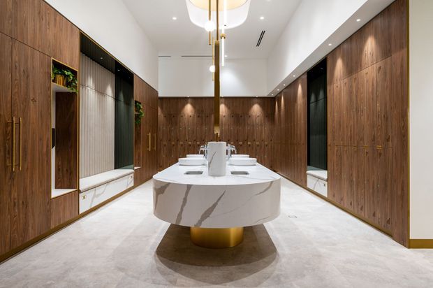 Luxurious contemporary walk-in wardrobe or dressing room with extensive dark timber built-in cabinetry, integrated open shelving, mirrors, and a central oval marble-topped island with a brushed gold pedestal. The room has a light stone or concrete-look floor, white ceiling, recessed spotlights, and a sculptural pendant light. Built-in display niches and greenery add detail, while the symmetrical layout creates a refined, hotel-like atmosphere.