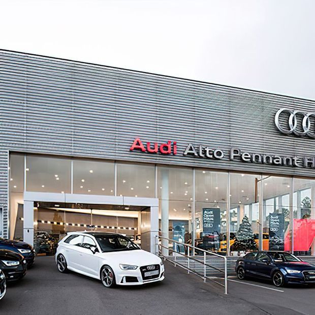 Exterior view of an Audi car dealership with a contemporary showroom facade. The building features extensive floor-to-ceiling glazing, horizontal metallic cladding, illuminated signage, entrance doors, accessibility railings, and vehicles displayed in front of the showroom.
