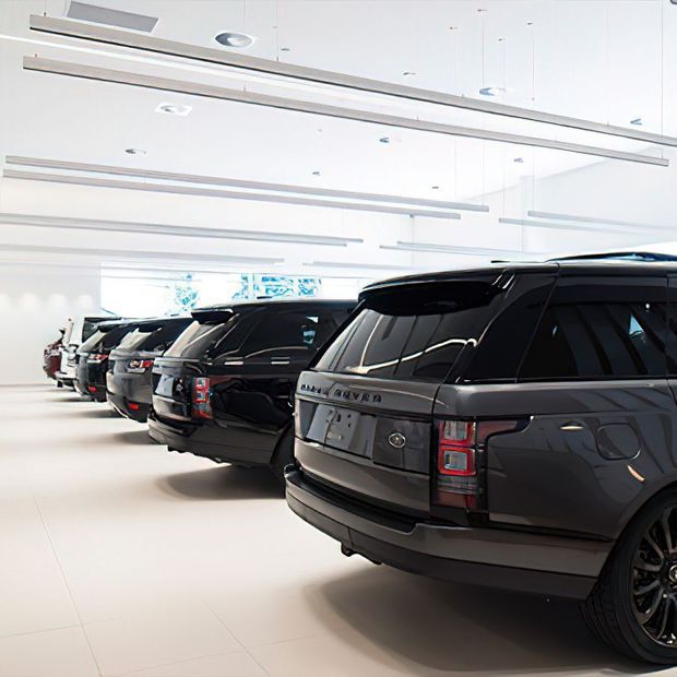 Bright, spacious indoor vehicle showroom or display garage with a row of dark luxury SUVs parked diagonally on a pale tiled floor. The space has white walls and ceiling, extensive linear recessed and suspended lighting, and large bright glazing or display panels in the background.
