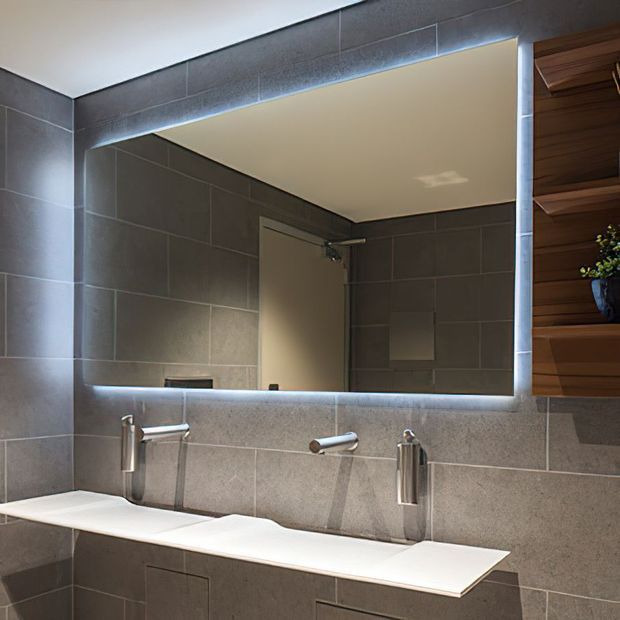 Contemporary bathroom vanity with a wide backlit rectangular mirror, double wall-mounted chrome faucets, a long white benchtop, and dark grey tiled walls. A timber slatted built-in shelving feature is visible at the right edge.