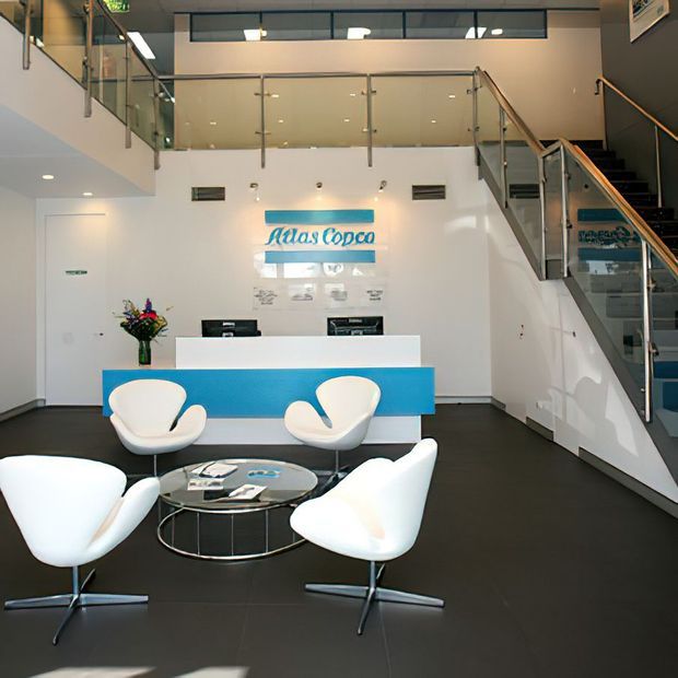 A bright, contemporary commercial reception and waiting area with a white reception desk featuring a turquoise-blue front panel, Atlas Copco branding, modern white lounge chairs, and a round glass coffee table. The space has dark flooring, white walls, glass balustrades, and a staircase leading to a mezzanine level.