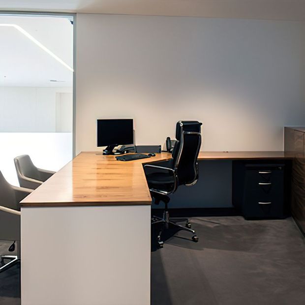A compact contemporary home office with an L-shaped built-in desk, pale timber work surface, dark cabinetry and drawers, black office chairs, computer equipment, and grey carpet. A glazed opening provides daylight from an adjacent space.