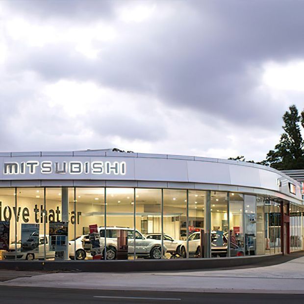 Exterior view of a modern Mitsubishi car dealership with a curved, glazed showroom facade, prominent illuminated signage, vehicles visible inside, and a broad paved frontage beneath an overcast sky.