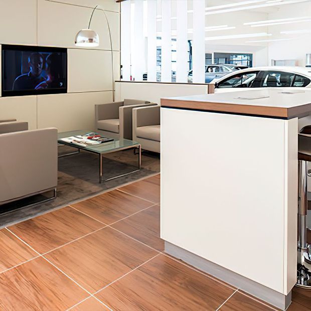 Bright contemporary automotive showroom or customer lounge with a white built-in reception or display counter, a compact seating area, wall-mounted television, pendant light, large windows, and vehicles visible beyond the glazing. The space has warm wood-look tiled flooring and clean white architectural finishes.