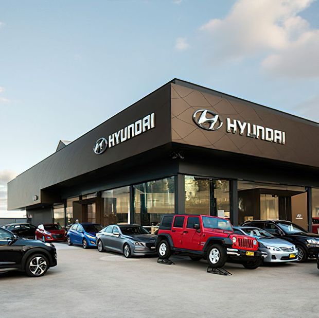 Exterior view of a modern Hyundai car dealership with a dark angular canopy, extensive floor-to-ceiling glazing, prominent illuminated Hyundai signage, and multiple vehicles parked across the forecourt.