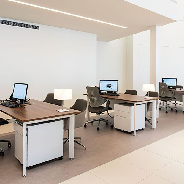 Bright, minimalist open-plan office with multiple wood-topped desks, white drawer units, ergonomic task chairs, computer monitors, and simple desk lamps. The space features white walls, pale flooring, recessed linear lighting, and a clean contemporary fit-out.