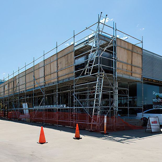 Exterior view of a large commercial or public building under construction, wrapped in extensive metal scaffolding and timber safety hoarding. Red construction fencing and orange traffic cones line the paved foreground, with a partially visible finished facade and signage behind the works.