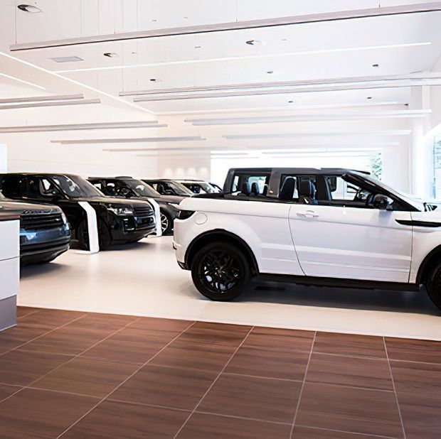 Bright, modern luxury car showroom displaying several SUVs, including a prominent white SUV in the foreground. The space has a highly illuminated white interior with layered linear ceiling lighting and large-format brown wood-look floor tiles.