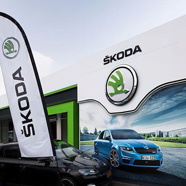 Exterior promotional image of a Škoda automotive dealership or showroom, with a white facade, large Škoda branding and circular logo, a branded feather flag, and Škoda vehicles displayed in front. Bright green and black facade accents are visible around the entrance.