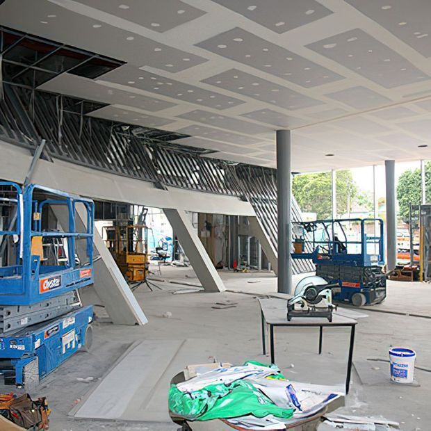 Interior construction photograph of a large, partially completed commercial or public building. The space has exposed structural elements, slanted supports, a dramatic metal balustrade, tall columns, and a patterned suspended ceiling with recessed lights. Construction equipment, tools, materials, and unfinished surfaces occupy the foreground and surrounding area.