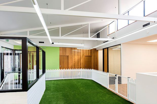 Bright contemporary interior with a large open atrium or indoor courtyard. Artificial grass covers the floor, bordered by white picket-style fencing and white railings. Black-framed glazed partitions, a vivid green wall, warm timber wall panelling, and exposed white structural ceiling beams define the space.