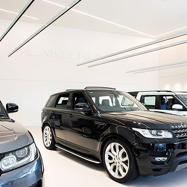 Bright, minimalist vehicle showroom displaying several luxury SUVs, dominated by a black Range Rover in the centre. The space has white wall and floor finishes, integrated linear ceiling lighting, and clean contemporary detailing.