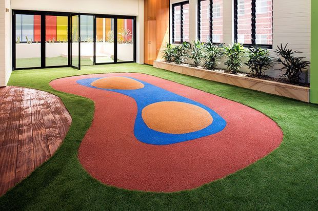 A vibrant indoor-outdoor recreation area featuring artificial grass flooring, a colorful red, blue, and orange rubber play surface with organic shapes, timber decking at the left, large black-framed glazed folding doors, and a planted raised garden bed beneath timber-lined walls and windows.