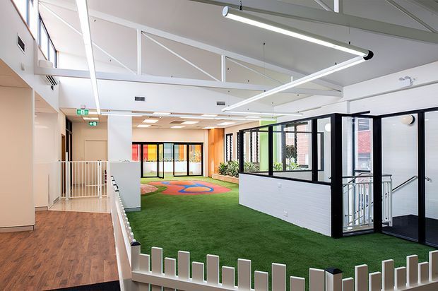 Bright contemporary indoor communal space with a large open play area covered in green artificial turf, colourful soft-play markings, black-framed glazed partitions, a small white picket-style barrier, and an adjacent timber-look corridor. The space has a high ceiling with exposed white structural beams, linear pendant lighting, skylights or roof glazing, and abundant natural light.