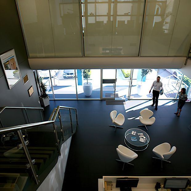 A high-angle view of a contemporary commercial lobby or reception area, featuring a dark floor, floor-to-ceiling glazing, a stairway with metal and glass balustrades, modern white lounge chairs around small round tables, and people near the entrance. A large internal glazed partition or mezzanine edge spans the upper portion of the image.