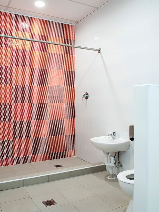 A compact commercial-style bathroom with an open shower area, white wall tiles, pale floor tiles, exposed plumbing beneath a wall-mounted basin, toilet, and a striking red, orange, and dark mosaic feature wall. The room has a suspended acoustic ceiling with recessed lighting and stainless-steel shower rail.