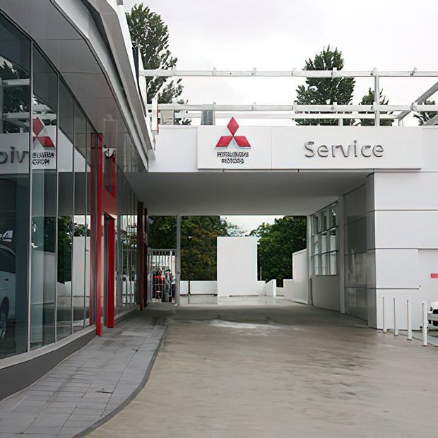 Exterior view of a modern Mitsubishi Motors automotive service facility. The image shows a covered vehicle service driveway between glazed showroom-style walls, with white panel cladding, red vertical accents, prominent Mitsubishi branding, and large 'Service' signage. Trees are visible beyond the building.