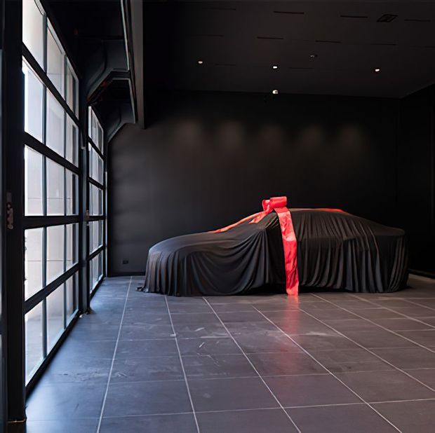 A dark, dramatic garage or showroom-like space with black walls and ceiling, large black-framed glazed doors along one side, and a vehicle fully covered by a black drape with a vivid red ribbon. The floor is finished in large grey tiles, and recessed ceiling lights provide subdued illumination.