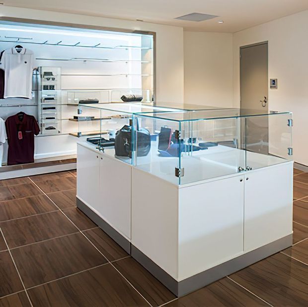 A bright, minimalist retail clothing boutique with white built-in wall shelving and hanging garment displays, a large central glass-topped display island with white cabinetry, and dark wood-look tiled flooring. The space has recessed lighting, clean lines, and a small staff or service door at the rear.