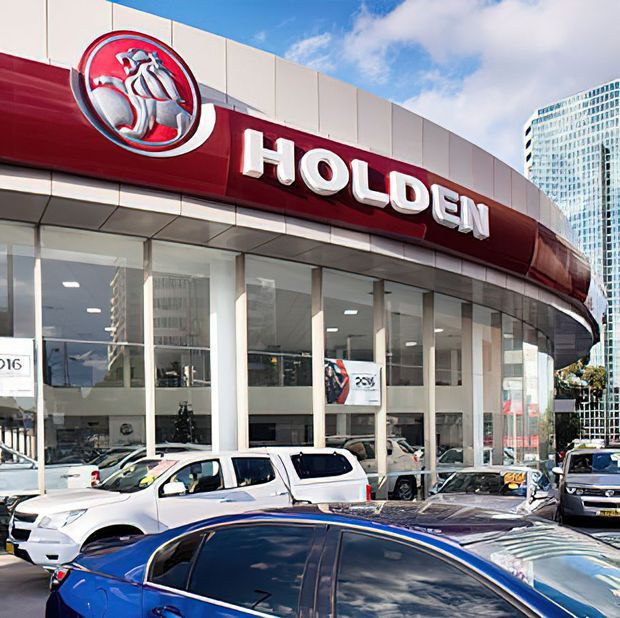 Exterior street-level view of a Holden automotive dealership, with a curved glazed showroom facade, prominent red Holden branding, displayed vehicles, and a modern high-rise building in the background.