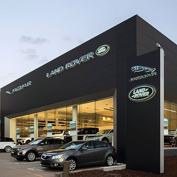 Exterior photograph of a contemporary Land Rover and Jaguar vehicle dealership. The building has a dark charcoal panelled facade, large floor-to-ceiling glazed showroom windows, illuminated automotive signage, and several vehicles displayed outside.
