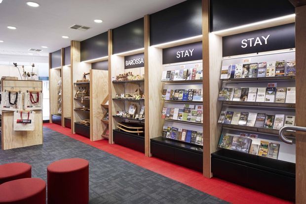 Interior view of a contemporary commercial visitor centre or retail information space, with built-in timber-framed display shelving and black backing panels filled with brochures and publications. Illuminated signage reads “STAY” and “BAROSSA.” The space has recessed ceiling lighting, dark patterned carpet, red carpet borders, and red upholstered cylindrical seating.