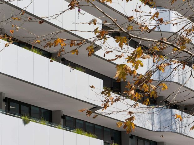 Modern multi-storey residential building viewed from outside, featuring crisp white panelled balcony fascias, dark recessed window and door frames, projecting slab-like balconies, and planted balcony edges. Bare and autumnal branches partially obscure the facade, while reflections appear in the glazing.