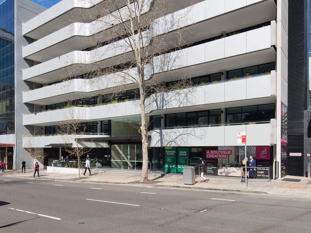 Street-level view of a contemporary multi-storey commercial building in an urban setting. The facade features broad white horizontal bands and projecting slab-like balconies or sunshades, dark recessed glazing, and a glazed ground-floor frontage with retail or commercial signage. Pedestrians and street trees line the pavement in front.