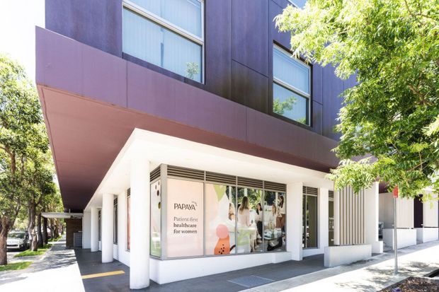 Street-facing view of a contemporary multi-storey commercial building with a projecting dark upper facade, white ground-floor storefront, large glazed windows, and slender vertical screening elements. The ground floor contains a healthcare retail tenancy with visible signage and display graphics, while mature street trees frame the frontage.
