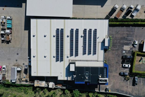 Aerial top-down view of a large commercial or industrial building with a light-coloured metal roof fitted with multiple long rows of solar panels and roof vents. Surrounding paved service areas contain parked trucks and cars, with landscaped vegetation along the perimeter.
