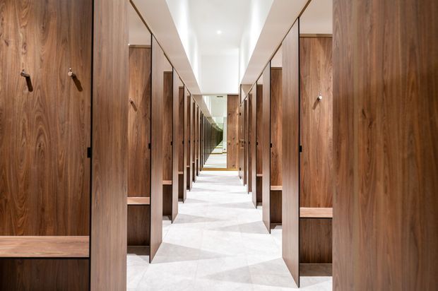 A long, symmetrical corridor lined on both sides with full-height dark timber storage or wardrobe joinery, each panel featuring hooks and open shelving. The space has a bright white ceiling with recessed downlights and a pale stone or terrazzo floor, creating a refined, highly ordered interior with strong perspective toward a distant glazed or open end.