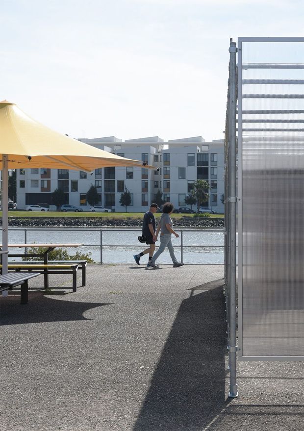 An outdoor waterfront promenade or courtyard beside a modern multi-storey apartment building. The paved space includes black picnic tables and benches beneath a large cream tensile shade canopy, with a translucent corrugated screen or windbreak forming a prominent vertical element at right. A metal railing borders the water, and two pedestrians walk along the path. The scene is bright, contemporary, and functional, with strong afternoon shadows.