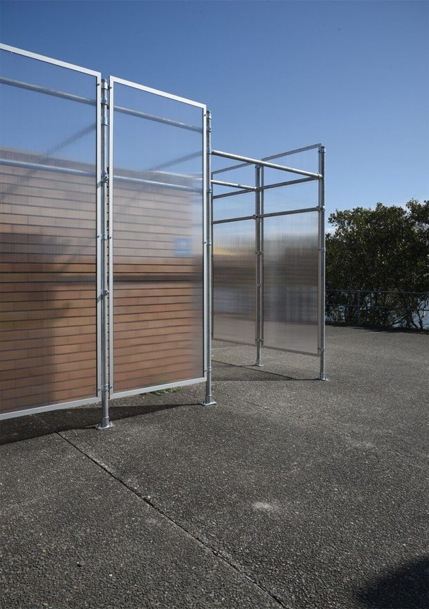 Exterior rooftop or elevated terrace featuring freestanding translucent corrugated screening panels in galvanized metal frames, arranged as a partially enclosed outdoor space. The panels provide privacy while allowing filtered light through, with a dark textured paved or asphalt-like surface and metal perimeter railing visible beyond. Clear blue sky and trees form the background.