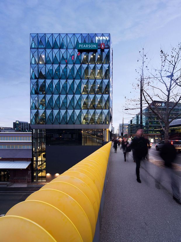 Twilight urban architectural photograph showing a contemporary multi-storey commercial office building with a striking blue glazed facade composed of repeated triangular geometric panels. The building is viewed from an elevated pedestrian walkway with a prominent bright yellow curved balustrade in the foreground. Adjacent city buildings, street activity, trees and pedestrians reinforce the dense urban setting.