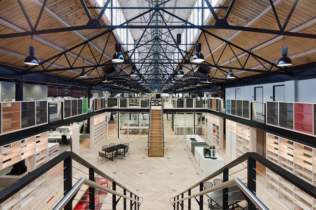 Wide elevated view of a large, open-plan commercial interior arranged around a central atrium. Black steel trusses and exposed services support a high timber-lined roof with a long skylight, while suspended black pendant lights illuminate display shelving, work areas, meeting tables, staircases and mezzanine-level joinery. The space has a contemporary industrial character with light herringbone flooring and extensive built-in cabinetry.