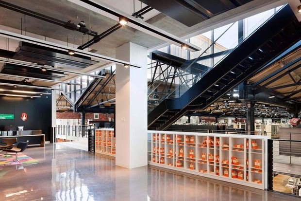 A spacious multi-level commercial interior with an industrial-style fit-out. The open space features exposed concrete ceilings and columns, visible structural steel, suspended black services and lighting, large glazed sections, a prominent black steel staircase, polished concrete flooring, and extensive white built-in shelving filled with orange-accented items. A lounge or reception area with seating is visible to the left.