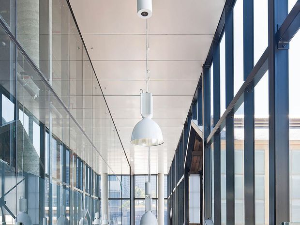 A bright, modern glazed corridor or internal passage with extensive floor-to-ceiling windows on both sides, dark metal window framing, a white suspended panel ceiling, exposed concrete columns, and a row of white pendant lights. Reflections in the glazing reinforce the open, airy commercial or institutional character.