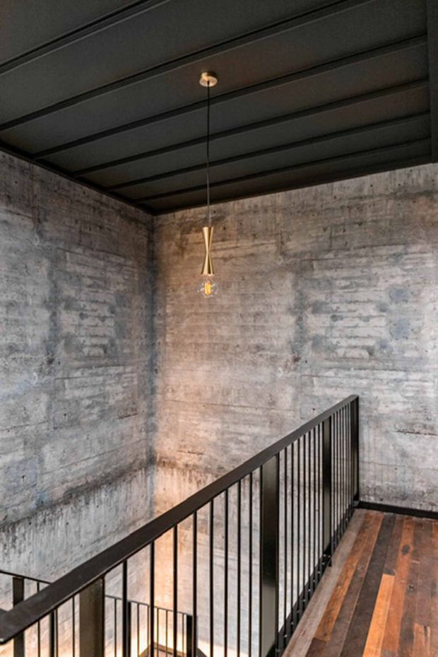 Industrial-style stair landing with exposed rough concrete walls, a dark ribbed metal ceiling, black metal balustrade, and mixed dark timber flooring. A minimalist brass pendant light hangs over the stairwell, creating a dramatic, moody interior.