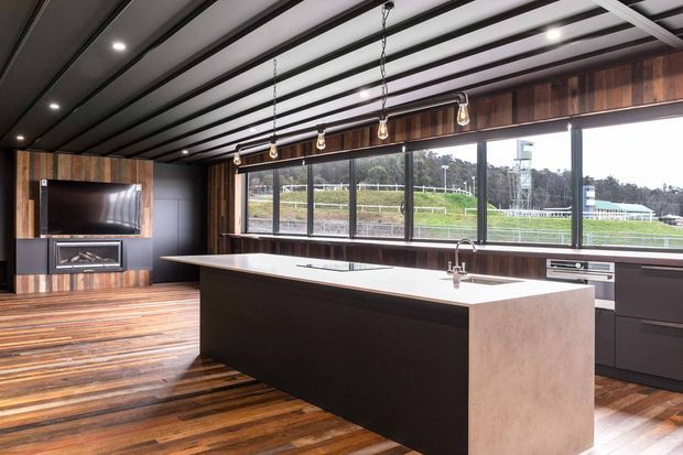 A contemporary open-plan kitchen and living area with a large central island, dark cabinetry, a pale stone benchtop, timber flooring, and broad black-framed windows overlooking a landscaped sporting or rural setting. The ceiling features exposed dark beams with recessed lighting, while a timber feature wall incorporates a television and fireplace.